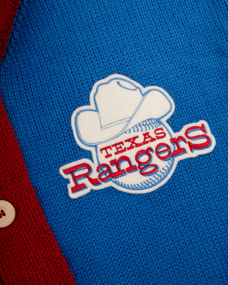 Close-up of the Texas Rangers 1972 Wool Shawl Collar Sweater by Ebbets Field Flannels, featuring a blue knit with the team’s white cowboy hat and baseball logo, red lettering, and a red border with white button in classic Replica MLB style.