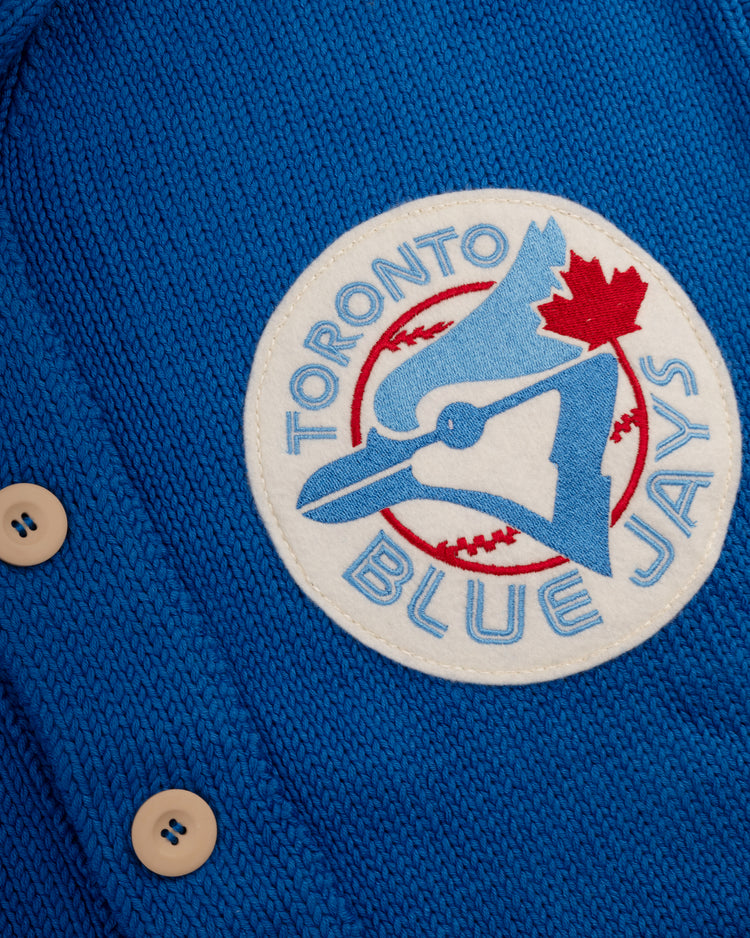 Close-up of the Ebbets Field Flannels Toronto Blue Jays 1977 Wool Shawl Collar Sweater in blue knit, with beige buttons and a round patch featuring a blue jay head, red maple leaf, and baseball background.