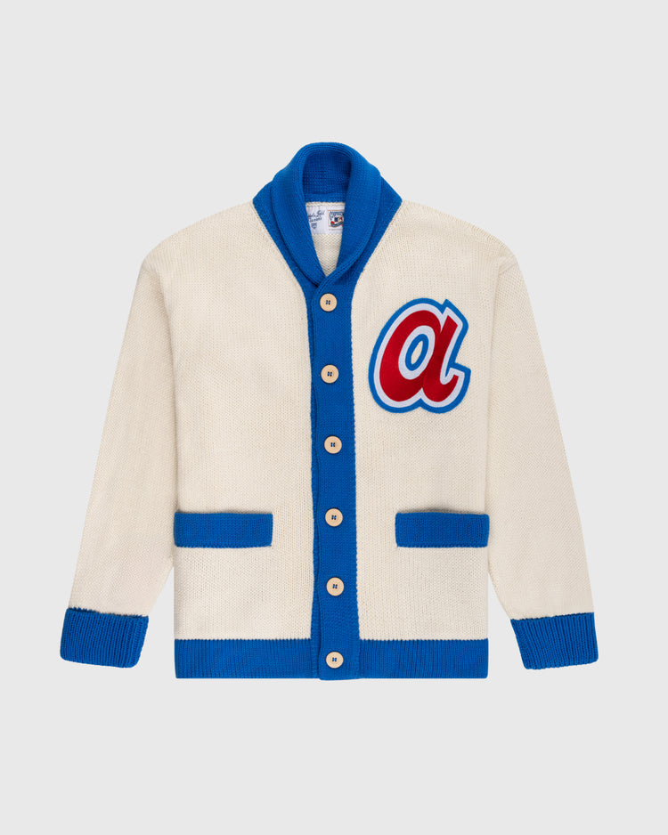 The Atlanta Braves 1974 Wool Shawl Collar Sweater by Ebbets Field Flannels is cream with blue trim, blue cuffs, two blue front pockets, tan buttons, and a large red and blue letter A patch inspired by vintage MLB and Braves styles.