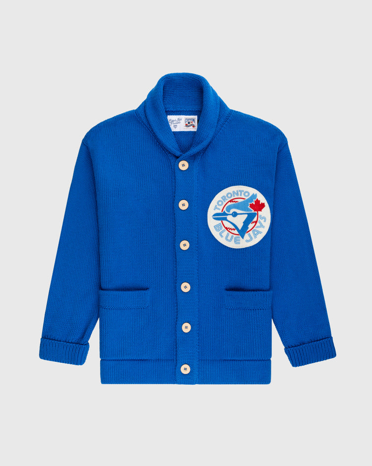 The Ebbets Field Flannels Toronto Blue Jays 1977 Wool Shawl Collar Sweater features royal blue knit, white buttons, two front pockets, and a large team logo patch on the left chest—styled after vintage on-field baseball sweaters.