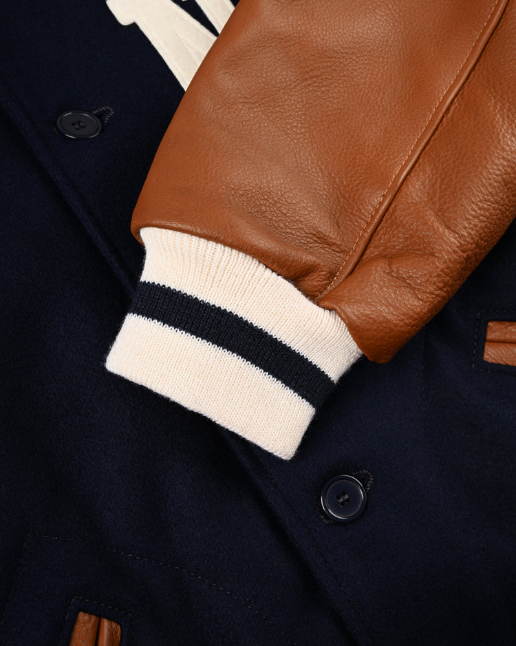 A close-up of the EFF New York Black Yankees 1940 Authentic Wool & Leather Varsity Jacket features brown leather sleeves, a white and navy striped knit cuff, navy wool body, black buttons, and a small brown leather accent.