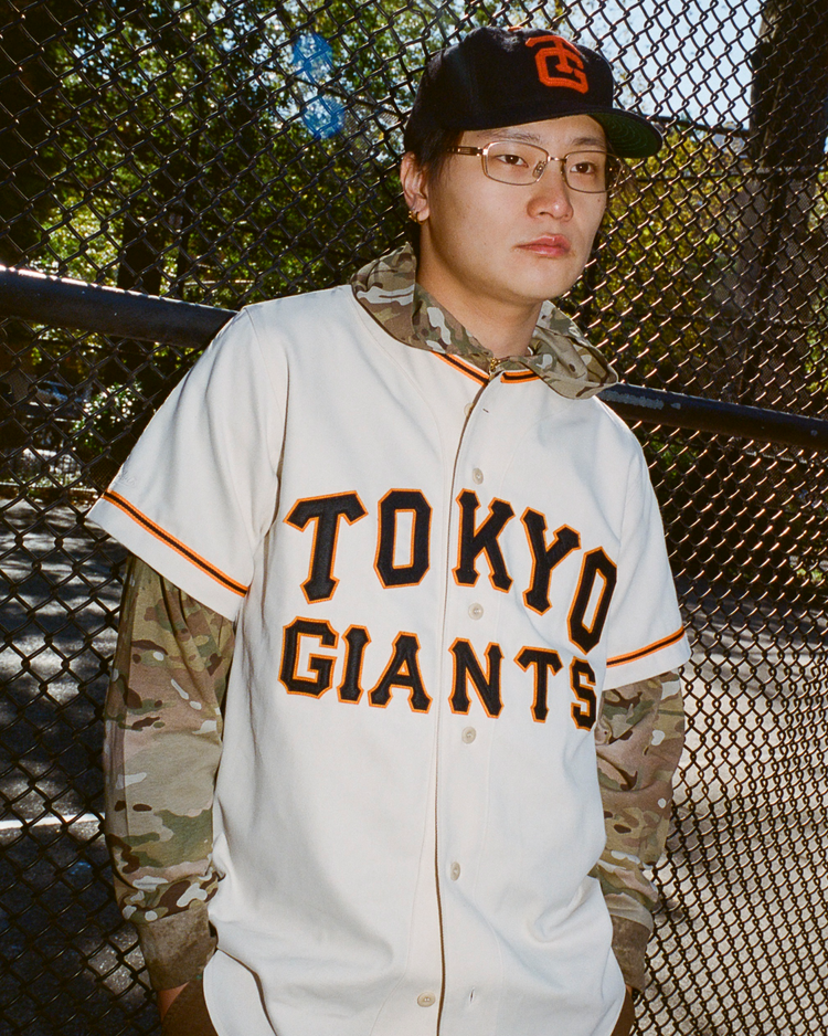 Person wearing a 'TOKYO GIANTS' baseball jersey and cap in front of a chain-link fence.