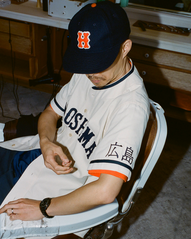 Person wearing a baseball cap and Hiroshima Carp jersey sitting on a chair indoors.