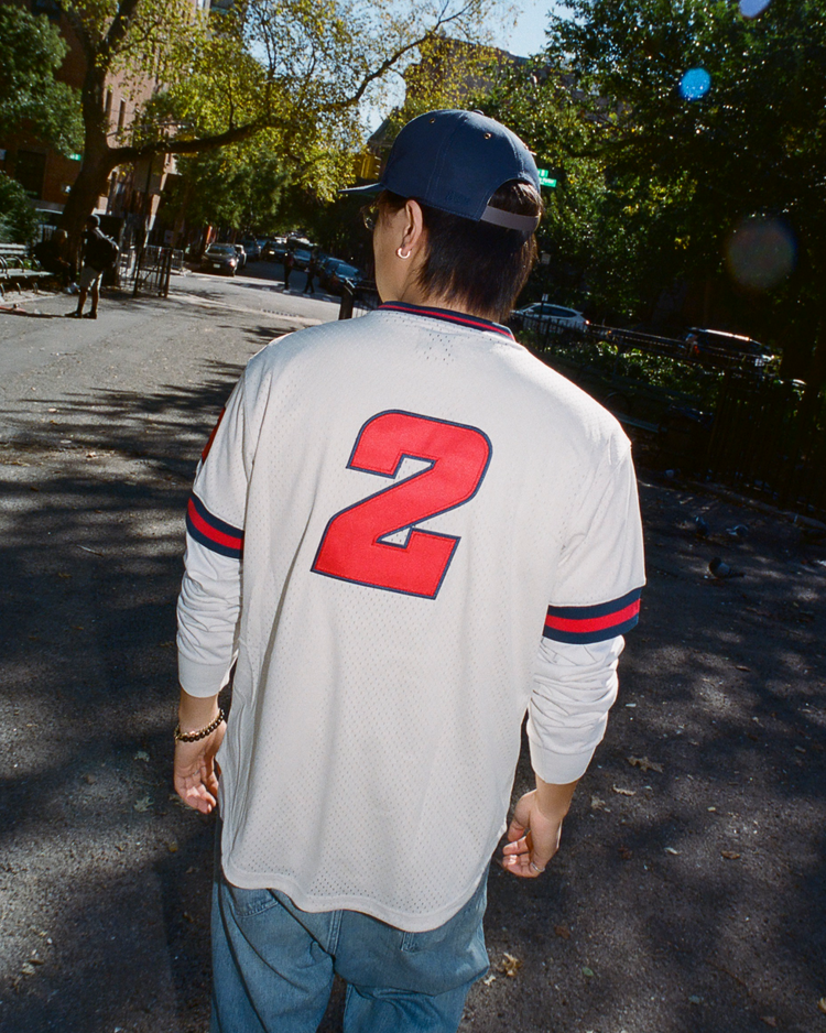 Person wearing a white Sankei Atoms mesh jersey with a red number 2 and blue cap walking on a street.