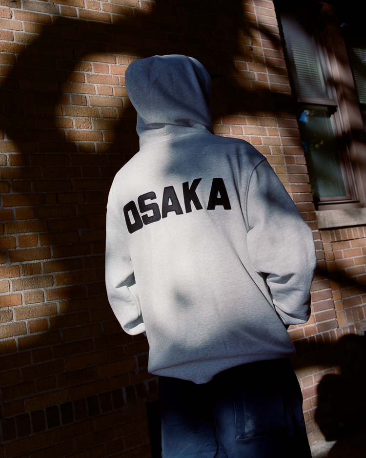 Person wearing a gray hoodie with 'OSAKA' printed on the back against a brick wall.
