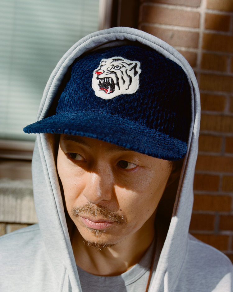Man wearing an indigo corduroy cap with an Osaka Tigers logo and gray hoodie against a brick wall.