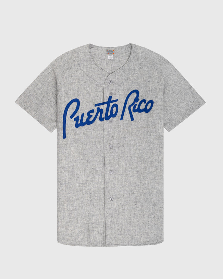 The Ebbets Field Puerto Rico 1958 National Team Authentic Wool Road Baseball Jersey is gray with short sleeves, blue Puerto Rico script on the chest, a button-up front, and a small neck tag.