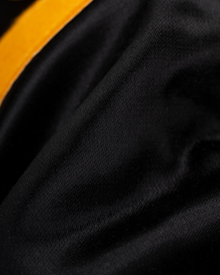 A close-up of smooth black satin fabric with a soft texture and a curved golden-yellow edge, from the EFF Roberto Clemente 1970 Authentic All Satin Varsity Jacket.