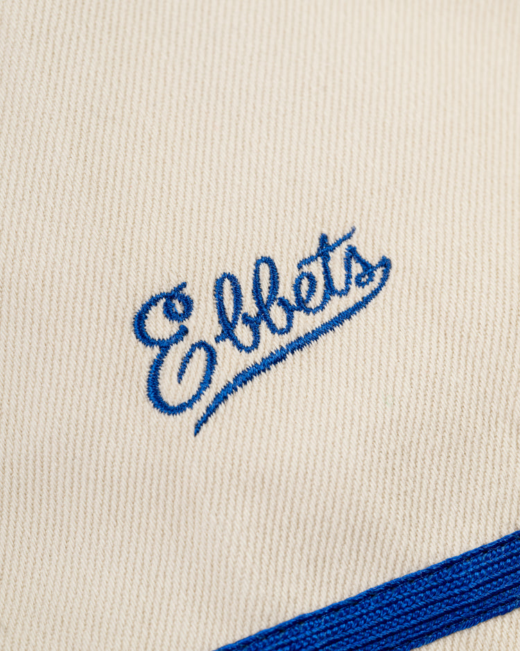 Close-up of cream-colored cotton twill from the Ebbets Field Santurce Cangrejeros Baseball Jersey, featuring Ebbets embroidered in blue cursive and a diagonal blue stripe, inspired by Puerto Rican baseball heritage.