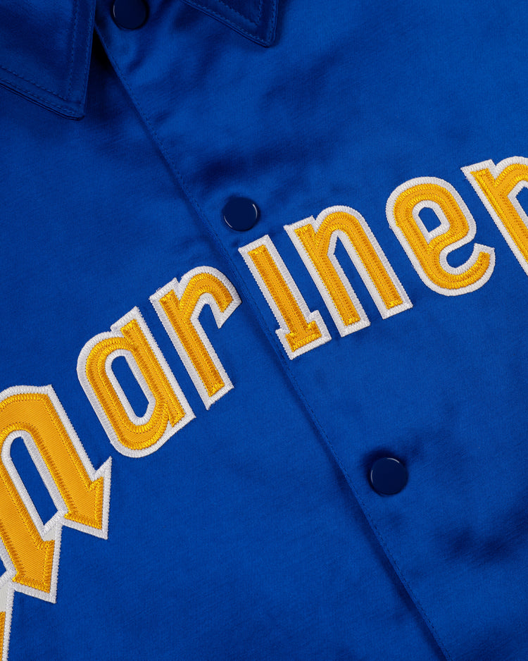 A close-up of the EFF Seattle Mariners 1982 Satin Windbreaker Jacket shows yellow and white mariner embroidery and dark blue snap buttons.