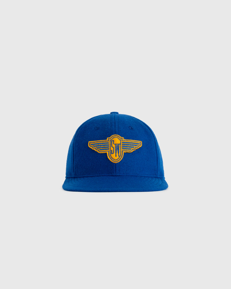 The EFF Stewart-Warner 1930 Authentic Wool Softbrim Ballcap is a blue cap with a flat brim, yellow and blue winged SN logo embroidery on the front, shown on a plain light gray background.