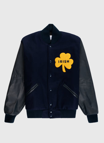 The EFF University of Notre Dame Authentic Wool & Leather Varsity Jacket features a navy body, black leather sleeves, black ribbed cuffs and collar, and a large yellow shamrock patch with “IRISH” on the chest.