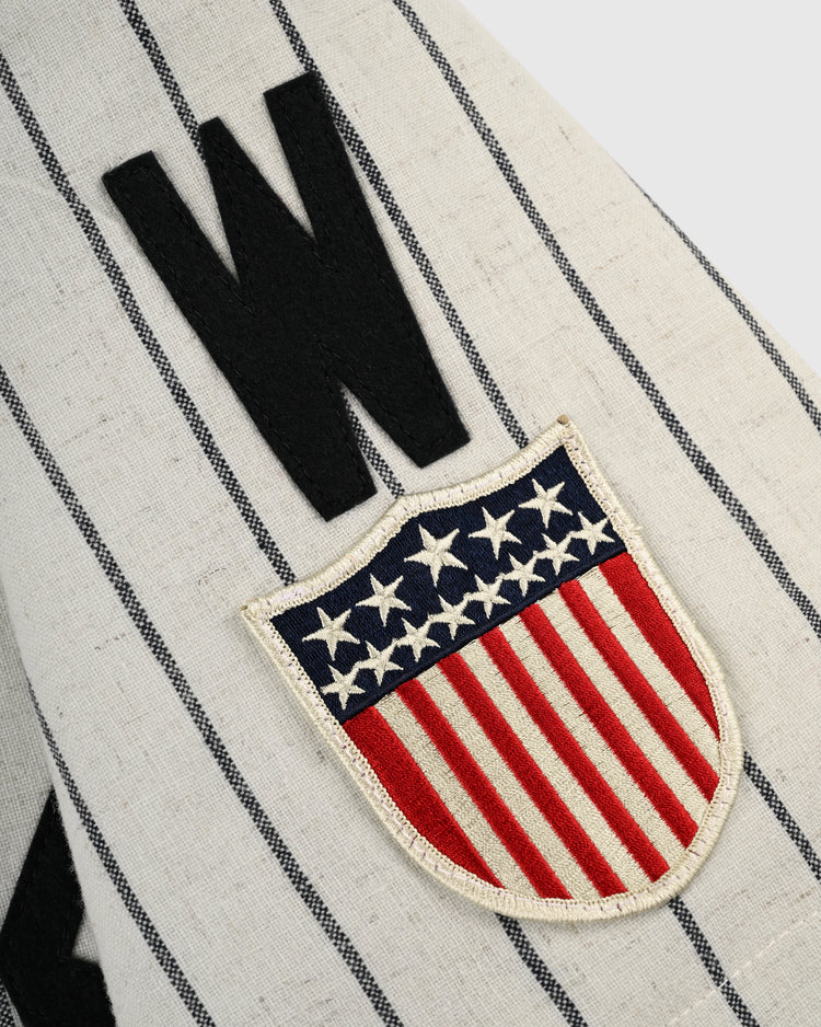 A close-up of the cream-colored, pinstriped Homestead Grays 1944 Authentic Wool Home Baseball Jersey by Ebbets Field Flannels, featuring a black W and a shield patch with stars and red stripes inspired by the American flag.