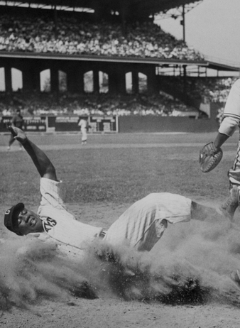 A baseball player slides into home plate as dust rises, while a catcher in uniform stands nearby watching the play. Spectators fill the stadium seats in the background.