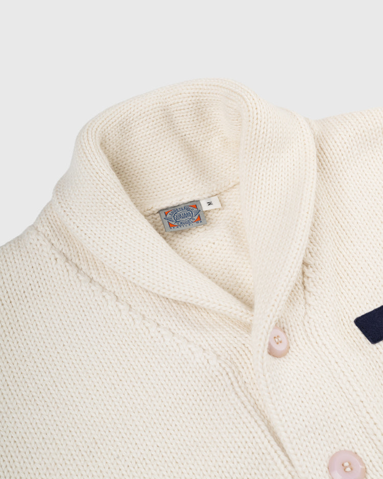 Close-up of the EFF Yale University 1923 Wool Shawl Collar Sweater, featuring a cream hue, light pink buttons, and brand label at the neckline. Its thick, textured knit recalls classic Yale Football traditions.