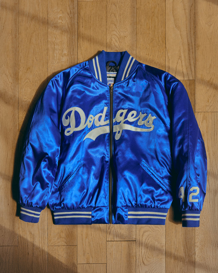 An EFF Brooklyn Dodgers 1947-50 Authentic All Satin Varsity Jacket in royal blue with white and gold lettering, striped collar and cuffs, and the number 42 on the left sleeve is laid flat on a wooden floor.