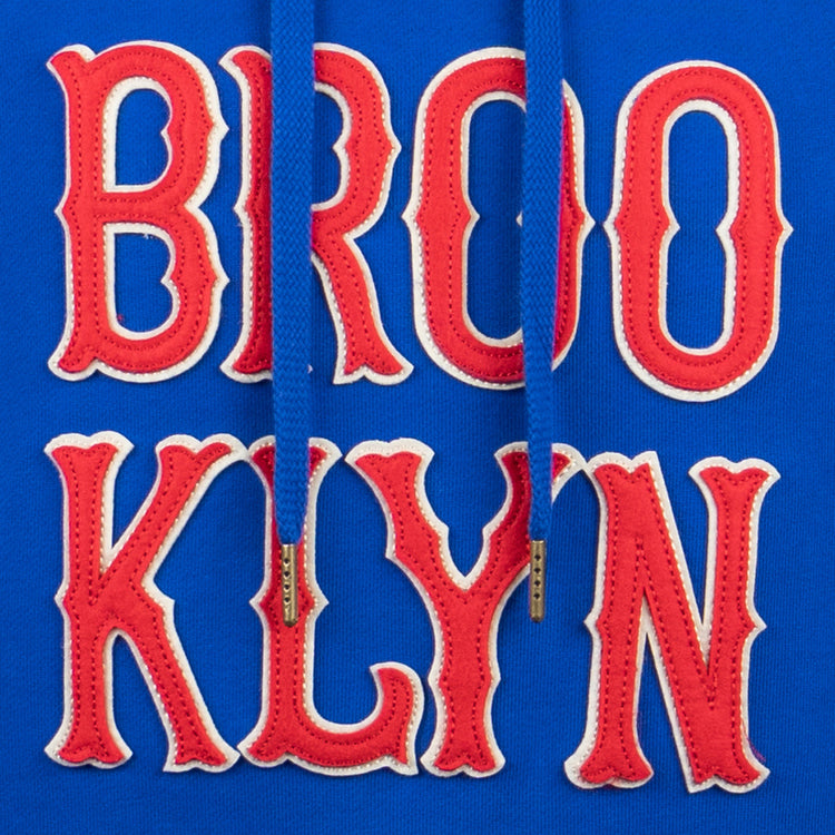 Close-up of EFF Brooklyn Royal Giants French Terry Script Hooded Sweatshirt: blue fabric with red and white BROO KLYN text, plus a matching blue drawstring with metal tips down the center.