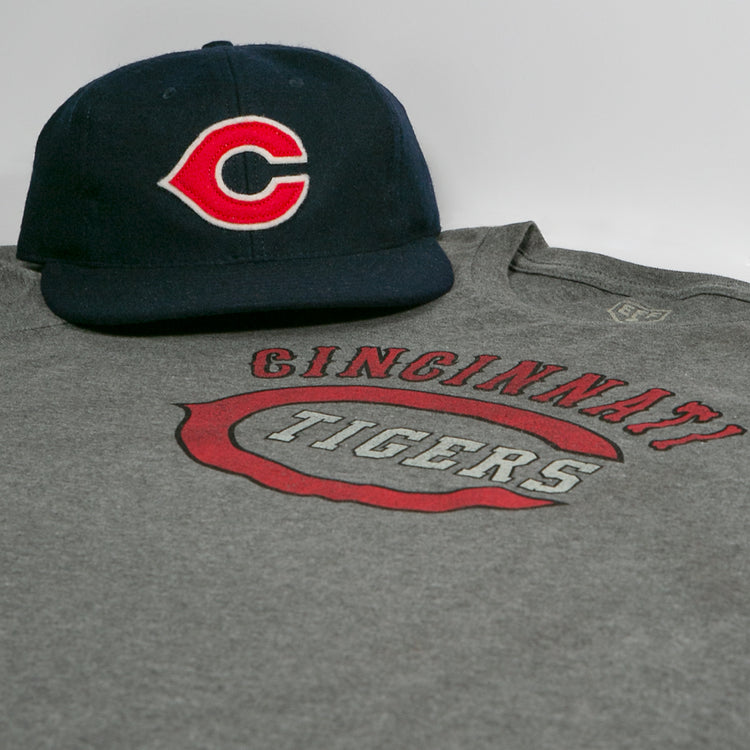 The EFF Cincinnati Tigers 1934 T-Shirt and a navy blue cap with a red and white C are displayed on a white surface, both inspired by the Negro American League.