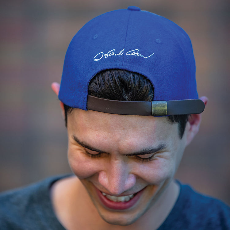 A smiling person wears the Ebbets Field Flannels Hank Aaron 1973 Signature Series Ballcap backwards, revealing white cursive writing on the back as they look down, with a blurred background behind them.