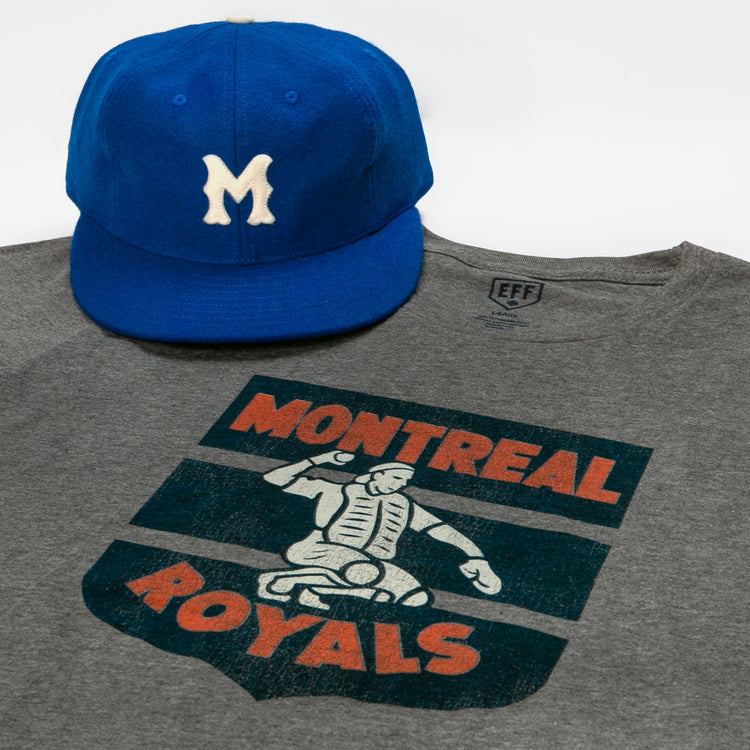 A blue baseball cap with a white M rests atop a gray EFF Montreal Royals 1946 T-Shirt, featuring a vintage-style catcher graphic and Dodger prospect roots.