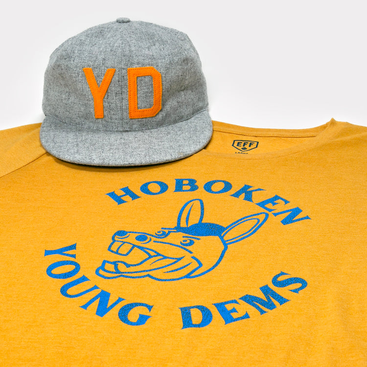 A gray cap with orange YD sits on a yellow Hoboken Young Democrats T-Shirt by Ebbets Field Flannels, featuring a blue cartoon donkey and Hoboken Young Dems in a circular design.