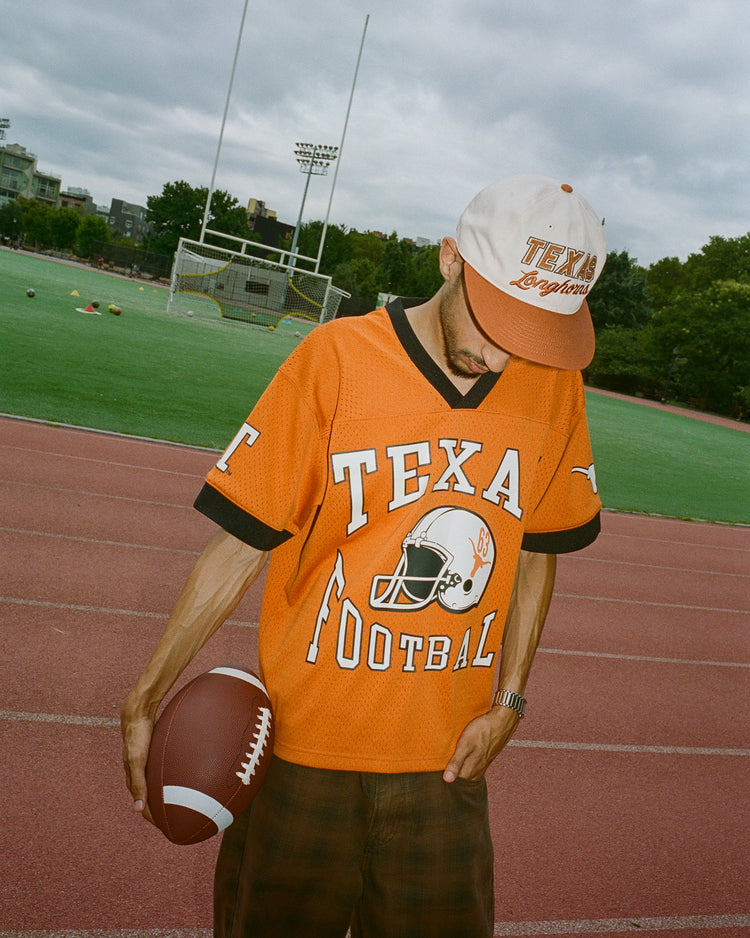 Wearing the Ebbets Field University of Texas Mesh Football Jersey and a matching cap, an individual stands on a running track holding a football. Behind them, the football field and goalposts are visible beneath a cloudy sky.