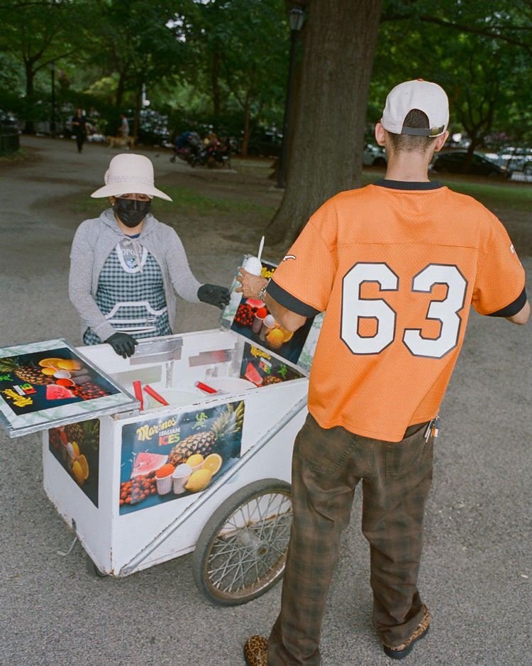 Wearing an Ebbets Field University of Texas Mesh Football Jersey, a person purchases a treat from a street vendor in a white hat and gloves, who stands behind an ice cream cart in the park.