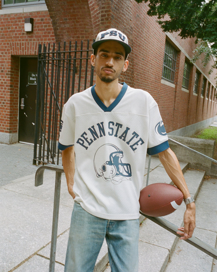 Wearing the Ebbets Field Penn State University Mesh Football Jersey and a matching cap, a person stands outside a brick building, holding a football under one arm and leaning on a railing.