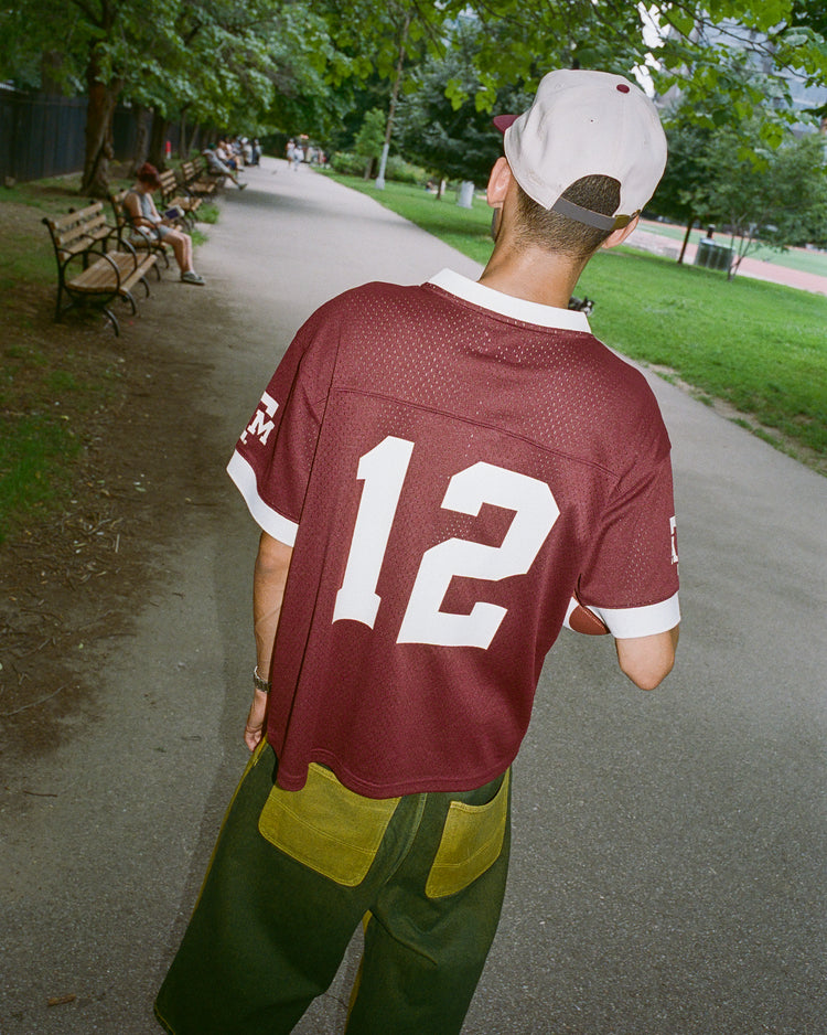 Wearing an Ebbets Field Texas A&M Mesh Football Jersey displaying the number 12, along with green shorts and a white cap, a person walks along a park path lined with benches and trees.