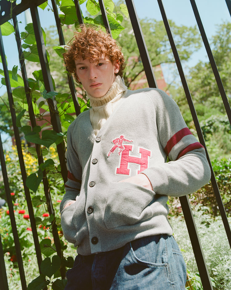 A young person with curly hair stands outdoors by a black fence, wearing the Ebbets Field Harvard University Cotton Varsity Cardigan—a gray cardigan featuring a red H and stripes—layered over a cream turtleneck. Green plants and trees fill the background.