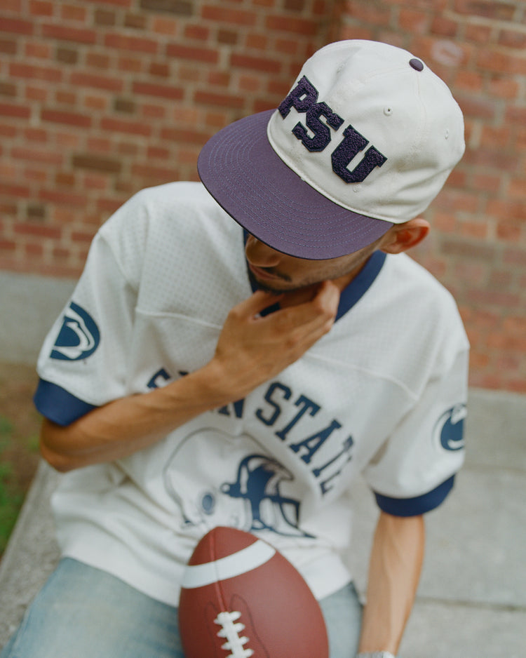 A person wearing a Penn State jersey and an Ebbets Field Penn State Chenille Logo Cotton Twill Ballcap sits outdoors holding a football, with a brick wall in the background. The cap, made from soft cotton twill, adds comfort and classic style to their look.