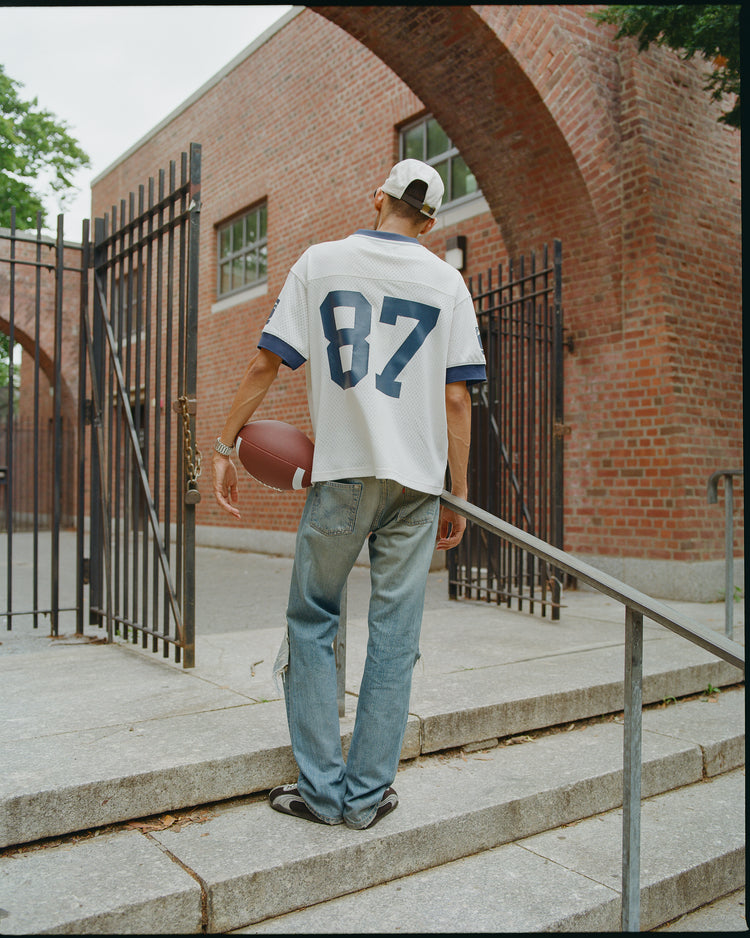 A person in a white cap, Ebbets Field Penn State University Mesh Football Jersey, and jeans stands by a metal railing, holding a football and facing away from the camera near a brick building and black iron gates.