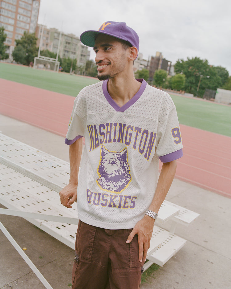 Wearing an Ebbets Field University of Washington Mesh Football Jersey and matching cap, a man stands by a white bench on the sports field, beaming. Trees and buildings in the background evoke the spirited atmosphere of a classic Rose Bowl game day.