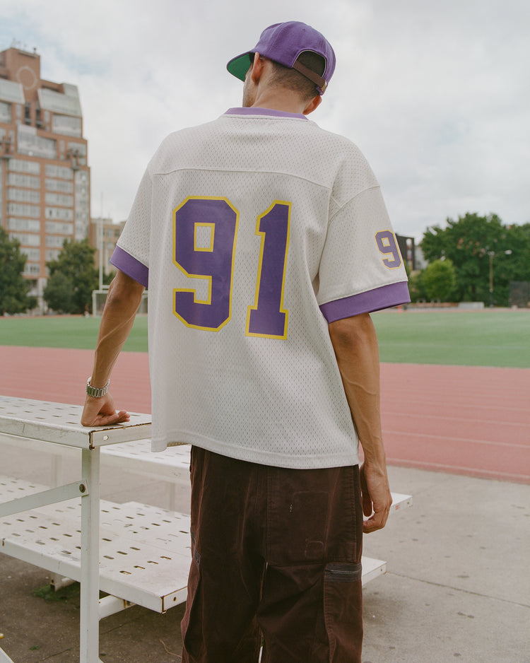 Wearing the Ebbets Field University of Washington Mesh Football Jersey—with purple and yellow 91—a person stands outdoors by a bench near a running track, facing away. They pair the jersey with a purple cap and brown pants, capturing the Huskies spirit. Buildings and trees appear in the background.