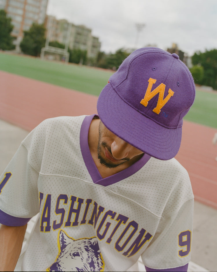 Wearing an Ebbets Field University of Washington Authentic Wool Ball Cap in purple, along with a white Huskies baseball jersey featuring the team’s wolf logo, a person stands near a track field and looks down, with trees and buildings visible in the background.