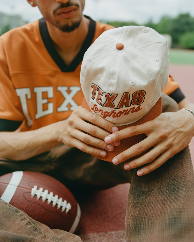 An individual wearing a Texas Longhorns jersey holds the University of Texas Longhorns Cotton Twill Ballcap by Ebbets Field, which features TEXAS Longhorns embroidery on the front. A football rests beside their leg on an outdoor sports field.