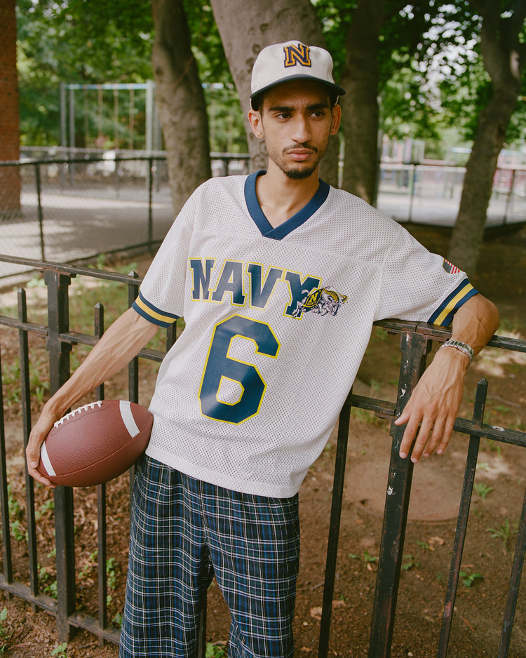 A person in a Navy 6 jersey and plaid pants holds a football and leans on a metal fence in a park. They wear the Ebbets Field Navy 1958 Logo Cotton Twill Ballcap, featuring an N for the US Naval Academy. Trees and a playground are visible in the background.