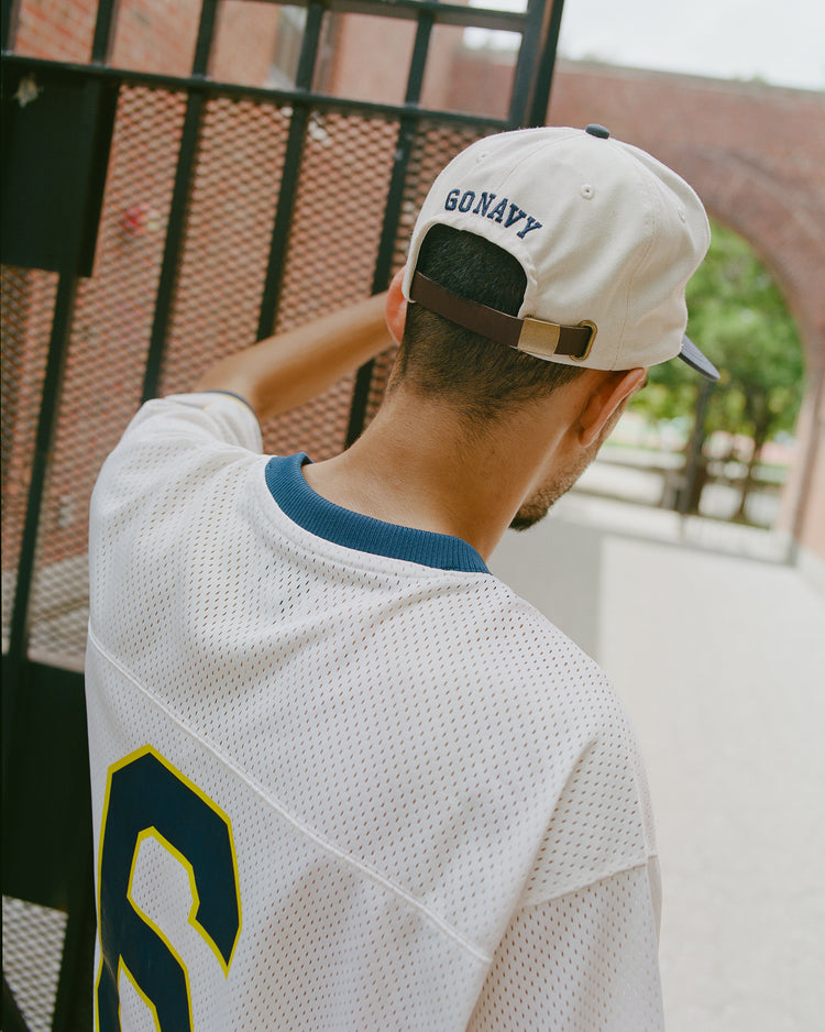 A person wearing a white mesh jersey with a blue and yellow 6 and an Ebbets Field Navy 1958 Logo Cotton Twill Ballcap stands by a metal gate, facing away from the camera—a scene reminiscent of Midshipmen football spirit at the US Naval Academy.