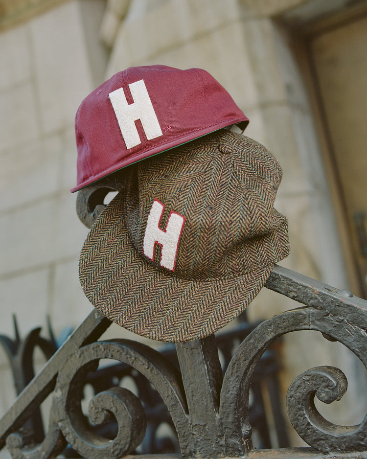 Two hats with prominent white H logos—a classic Harvard baseball cap and the Ebbets Field Harvard University Herringbone Wool Ballcap—rest on a decorative black iron fence in front of a stone building.