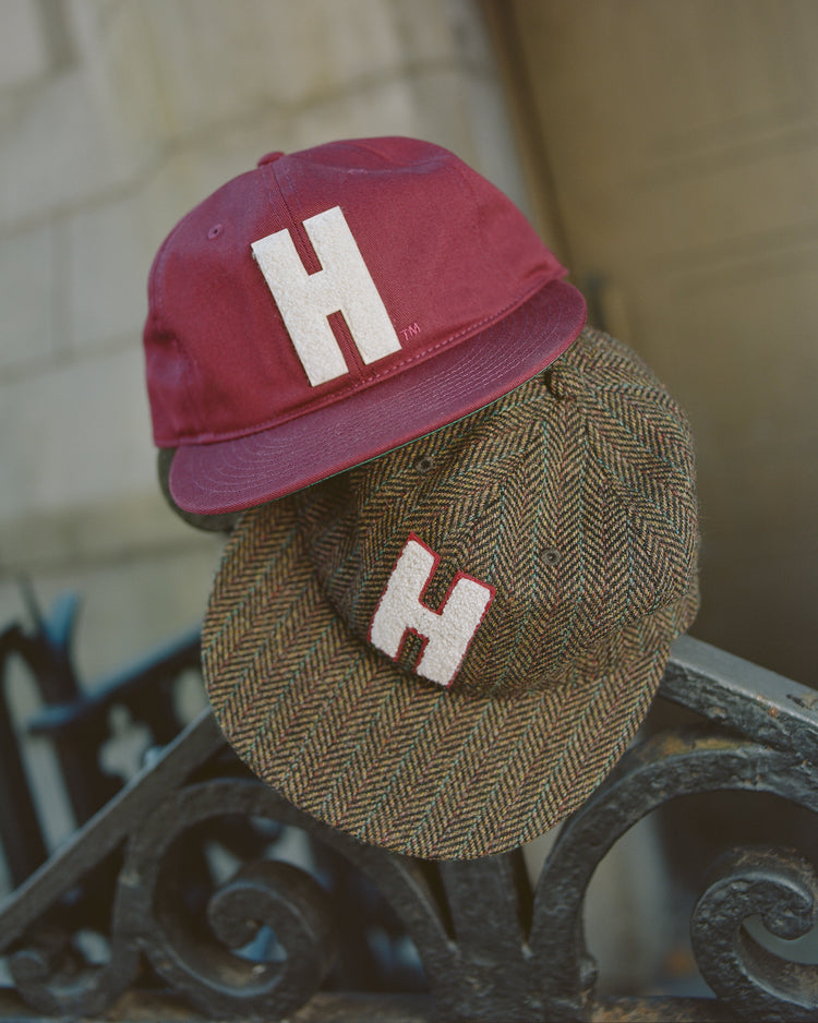 Two Harvard University Chenille H Cotton Twill Ballcaps by Ebbets Field, one in dark red and the other in a classic brown herringbone pattern, rest atop a black wrought iron fence. Each ballcap is crafted in traditional cotton twill and features a bold chenille “H” patch inspired by vintage Harvard baseball spirit, with an out-of-focus stone wall providing the backdrop.