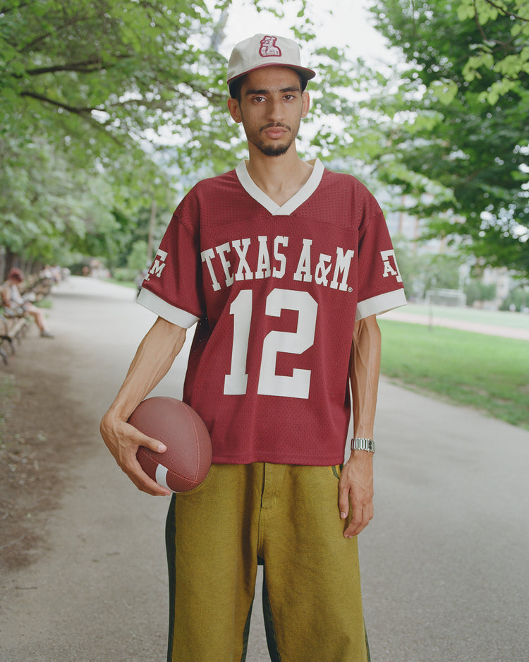 A man wearing the Ebbets Field Texas A&M Mesh Football Jersey and a matching cap holds a football in one hand, standing on a pathway lined with trees and benches in the background.