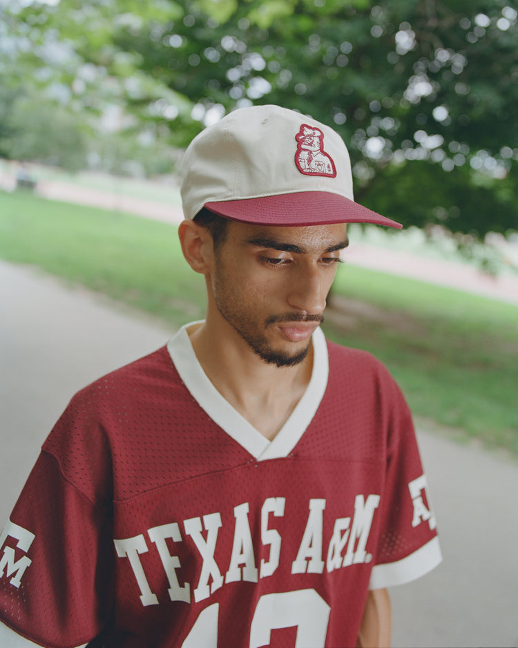 Wearing a maroon Texas A&M jersey and an Ebbets Field Texas A&M Sarge Cotton Twill Ballcap, a person stands outdoors on a path bordered by lush green trees and grass, embodying the tradition and spirit of Texas A&M history.
