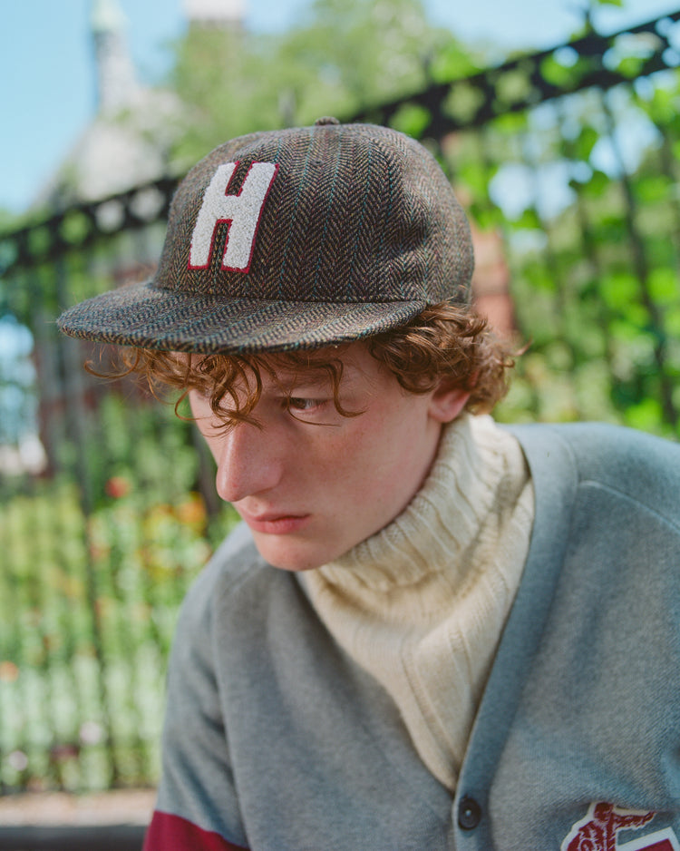 A young person with curly hair wears the Ebbets Field Harvard University Herringbone Wool Ballcap, along with a cream turtleneck sweater and a gray cardigan, sitting outdoors near a black metal fence and greenery.
