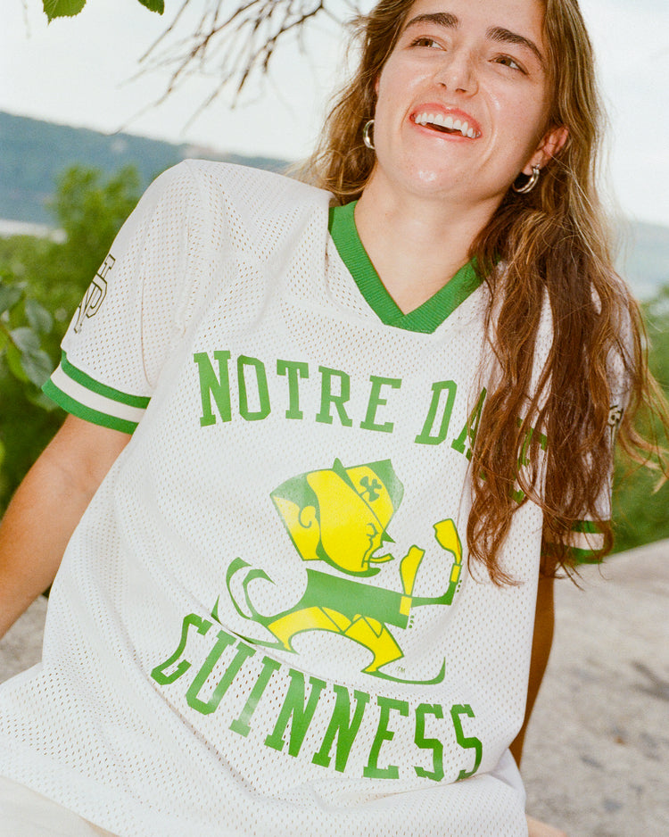 A smiling person with long brown hair is wearing the Ebbets Field Notre Dame X Guinness Mesh Football Jersey, sitting outdoors near greenery and rocks.