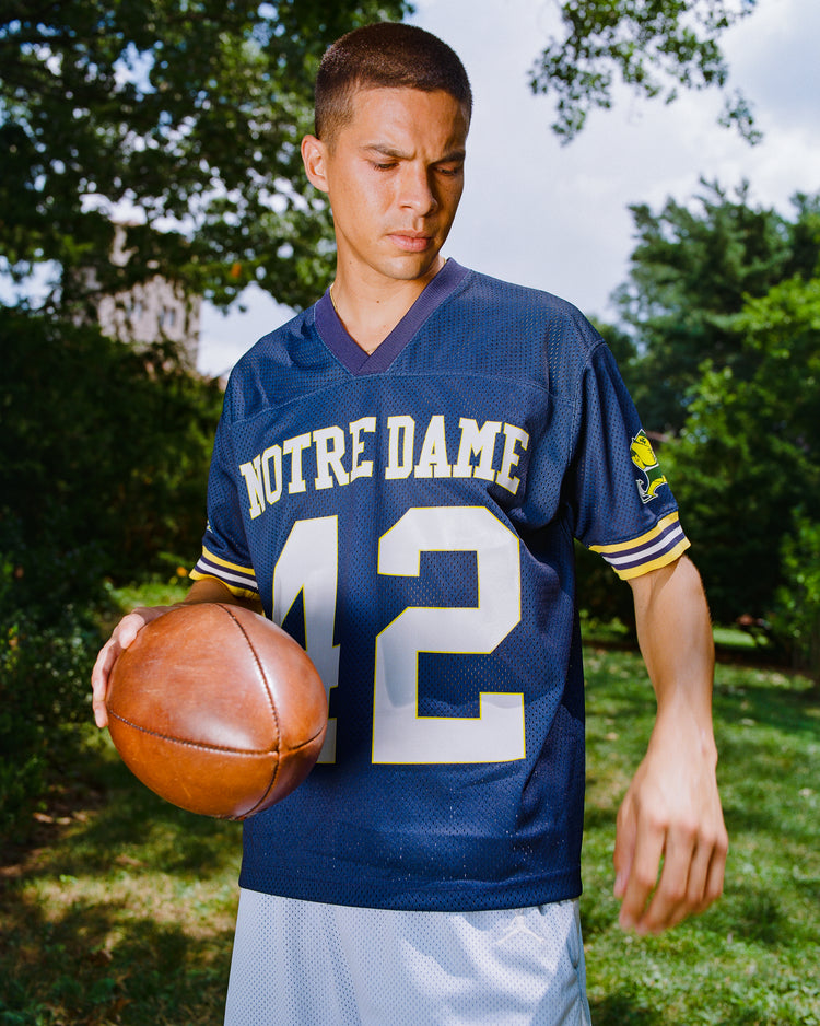 Wearing the Ebbets Field Notre Dame Mesh Football Jersey in blue, featuring the number 42, a person holds a classic brown football while standing on grass with trees behind them—perfectly capturing the enduring spirit of Fighting Irish football.