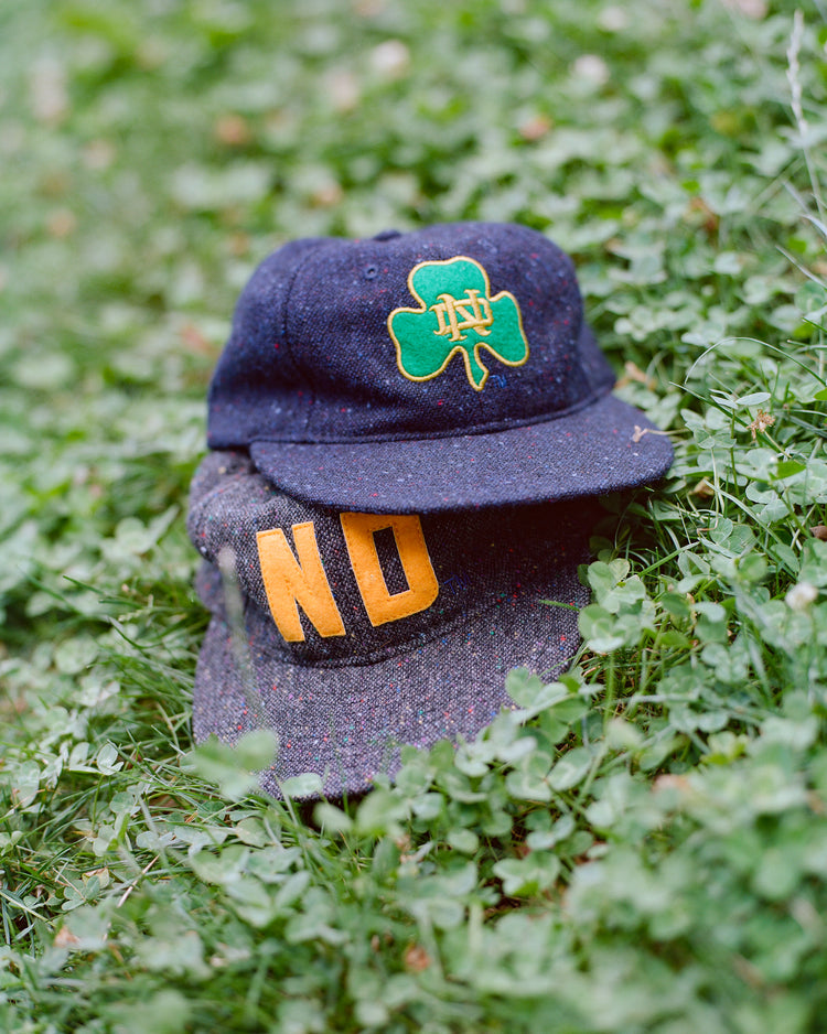 Two blue speckled Notre Dame X Magee Irish Donegal Wool Ballcaps by Ebbets Field, inspired by Irish heritage hats, rest on green clover. The top cap features a green shamrock with an ND logo, while the bottom cap displays large yellow letters spelling NO.