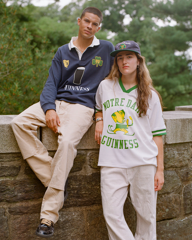 Two people in light khaki pants and casual Ebbets Field Notre Dame X Guinness Rugby Shirts lean against a stone wall outdoors, surrounded by green trees. One wears the classic navy rugby shirt, while the other sports the white version with a matching cap—both proudly celebrating their Irish heritage.