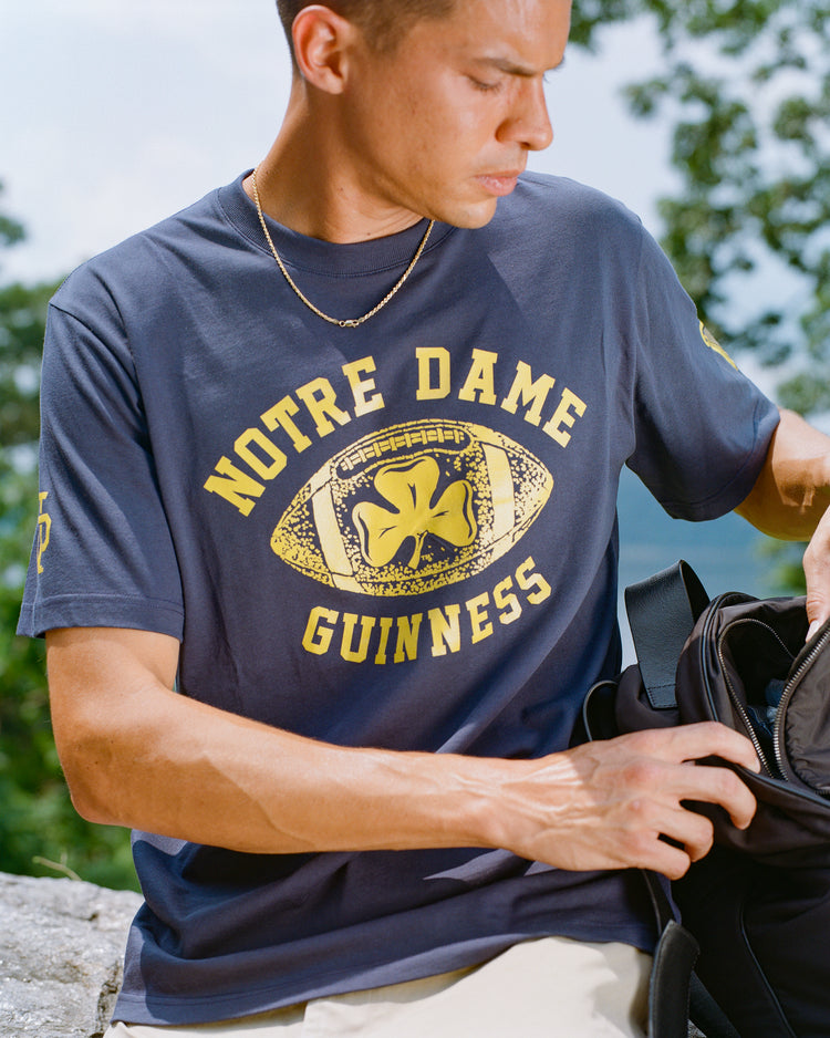 A person wearing the Ebbets Field Notre Dame X Guinness Short Sleeve Graphic Tee with a shamrock emblem looks down while reaching into a black bag outdoors.