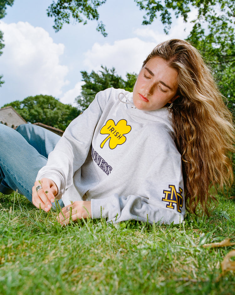 A person with long hair in a gray Ebbets Field Notre Dame X Guinness Heavyweight Pullover Hoodie sits on the grass, eyes closed, holding a blade of grass, with trees and sky in the background.
