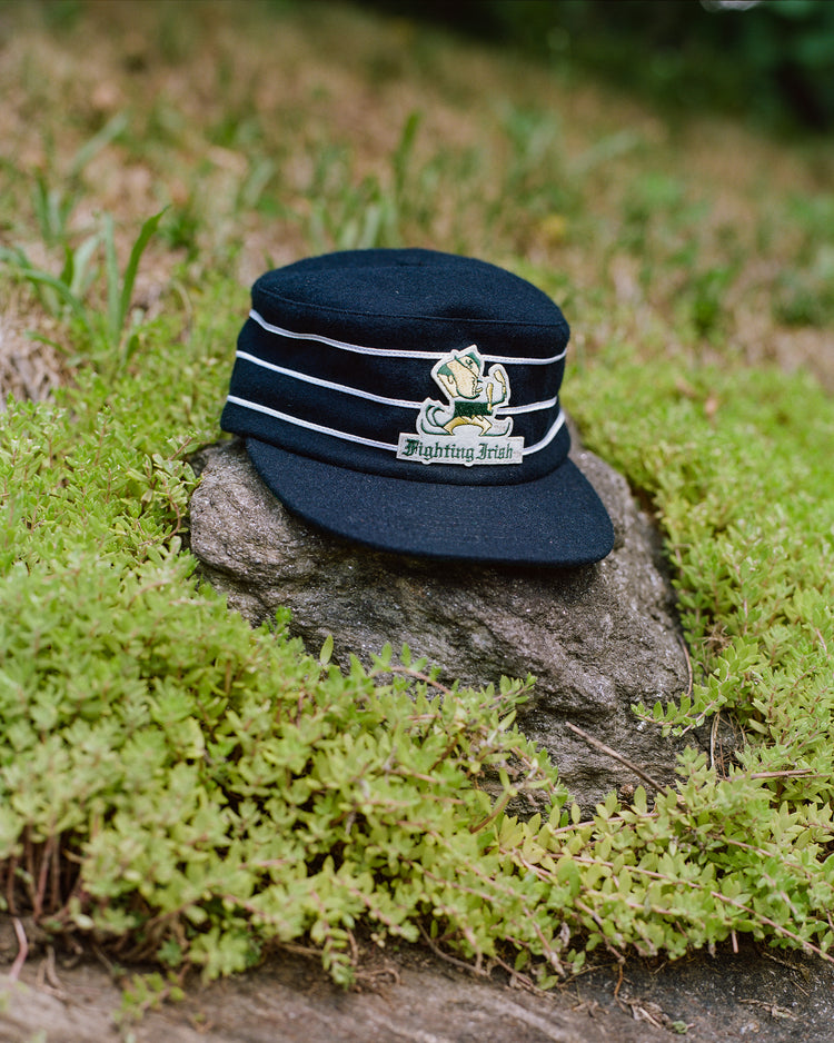 An Ebbets Field Notre Dame Leprechaun Pillbox Wool Ballcap sits on a rock amidst green grass and plants.